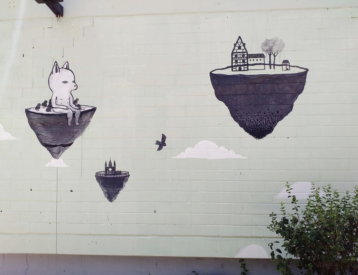 Mural of several floating rock islands painted in black and white on a pale green wall: one holds a large-eared creature with birds perched on it, another holds a half-timbered house and trees, a smaller one holds a church spire, with a bird flying between them.