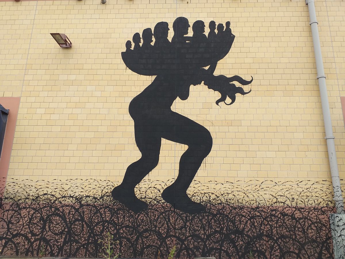 Black silhouette of a crouching nude figure carrying a cluster of smaller human silhouettes on its back like a bundle, wading through a field of barbed wire painted on a yellow brick wall.