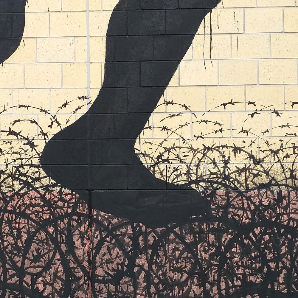 Close-up detail of a black silhouette leg and foot stepping through dense tangles of barbed wire, painted on a yellow brick wall.