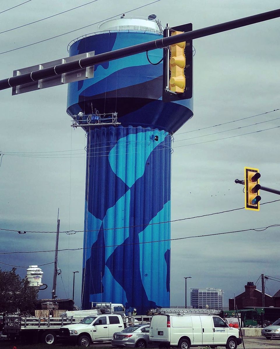 Water tower painted in dark blue camouflage, a worker standing on a suspended platform near the top, photographed through a traffic light and overhead cables against a grey sky.