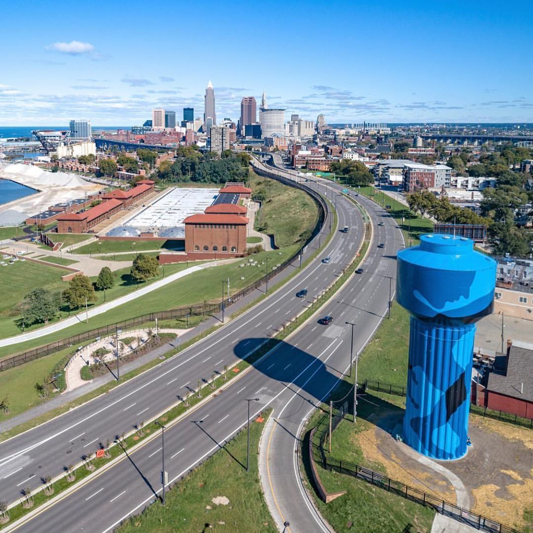 Aerial view of the camouflage-painted water tower beside a curving highway, with the Cleveland skyline and lake visible in the background under a clear blue sky.