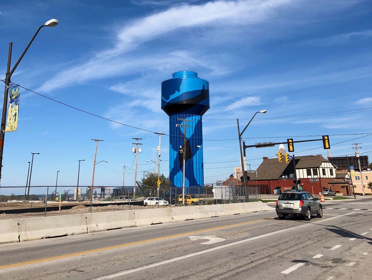 Wide street-level view of the blue camouflage water tower rising above a fenced lot, with a car driving past on the road and the city skyline faintly visible on the horizon.