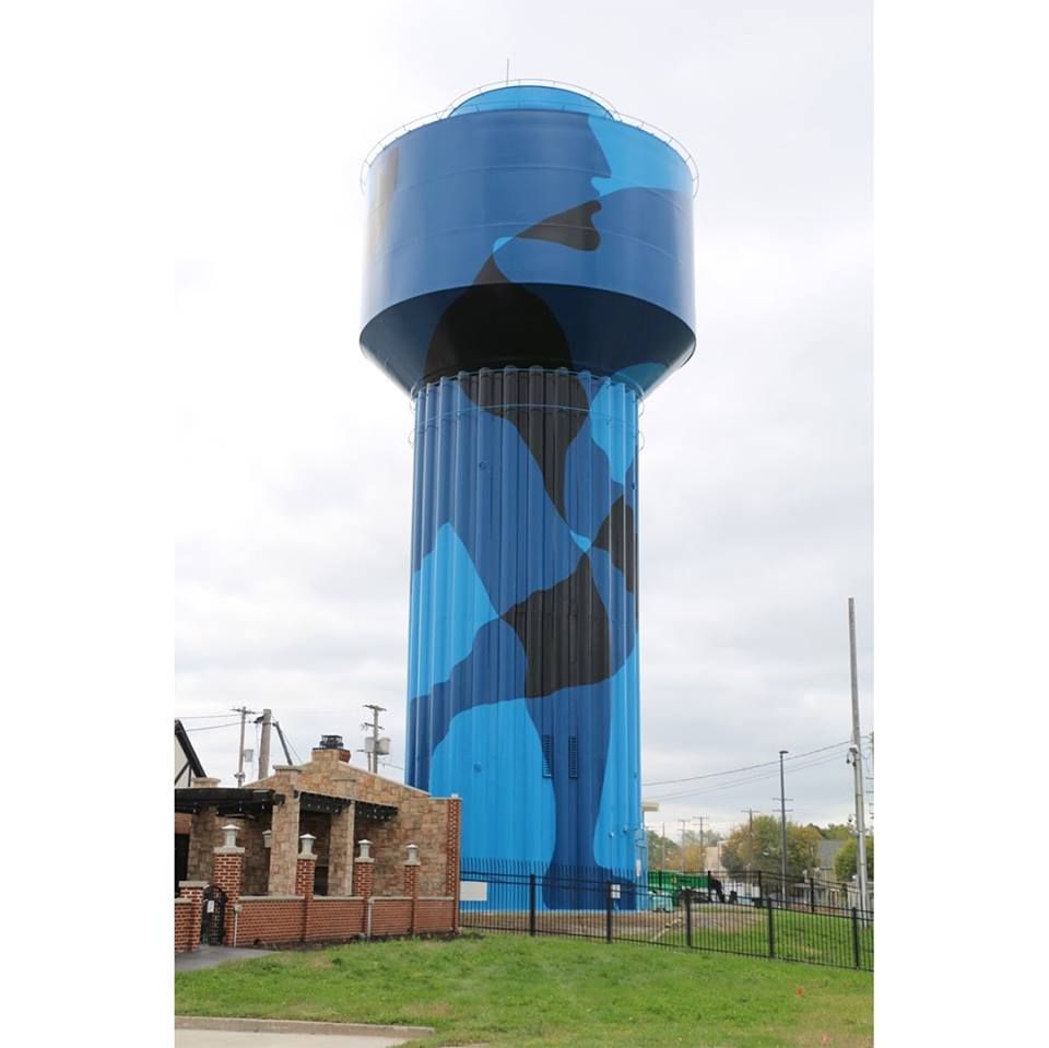 The completed water tower in blue and black camouflage, seen past a stone fireplace structure and brick wall under an overcast sky.