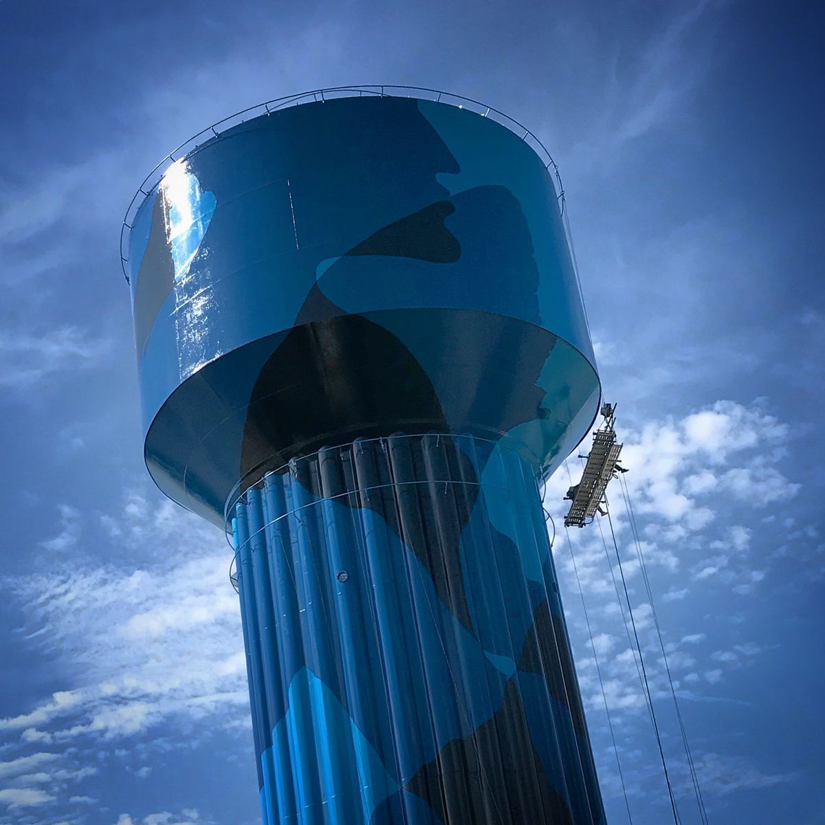 Low-angle view of the water tower's upper tank glinting in sunlight against a partly cloudy blue sky, with a suspended work platform hanging beside the fluted column below.