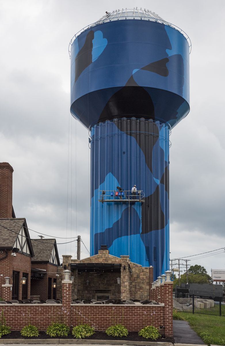 Historic cylindrical water tower painted in a dark blue and black camouflage pattern, with two workers on a suspended platform partway up the tank, seen behind a brick building and fenced yard.