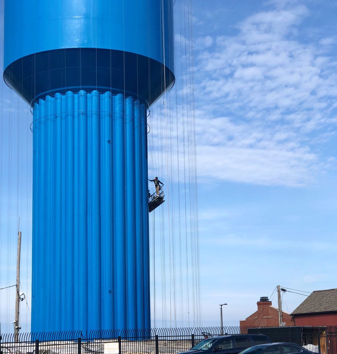 Close-up view of the water tower's fluted lower section painted solid blue, with a worker on a hanging platform against a partly cloudy sky, cars and a fence visible below.