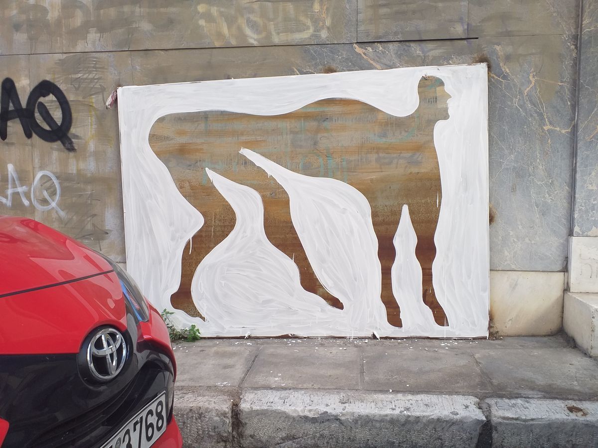 White painted silhouette of a bull-headed minotaur figure on a dark wood panel propped against a marble-clad wall, beside black spray-painted Greek tags and a parked red car.
