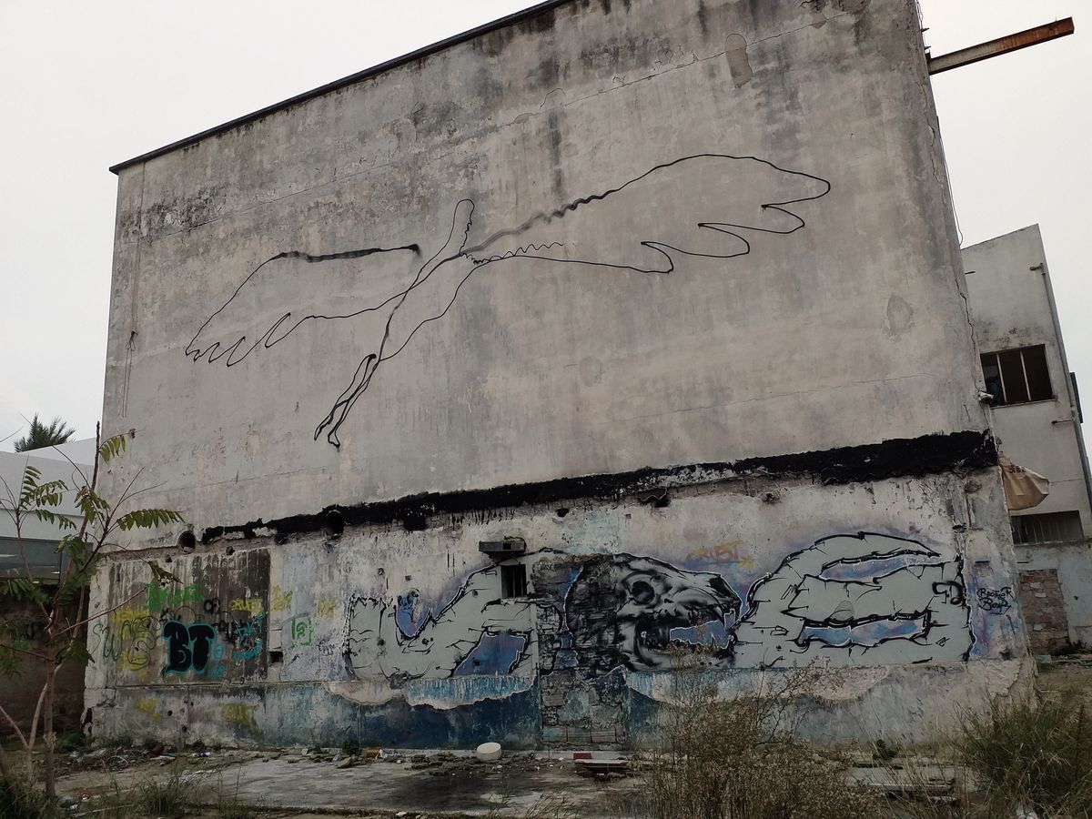 Thin black outline drawing of a falling or flying winged figure spanning a large concrete wall, above a black horizontal band and a weathered mural of a skull and abstract shapes on the lower section of an abandoned building.