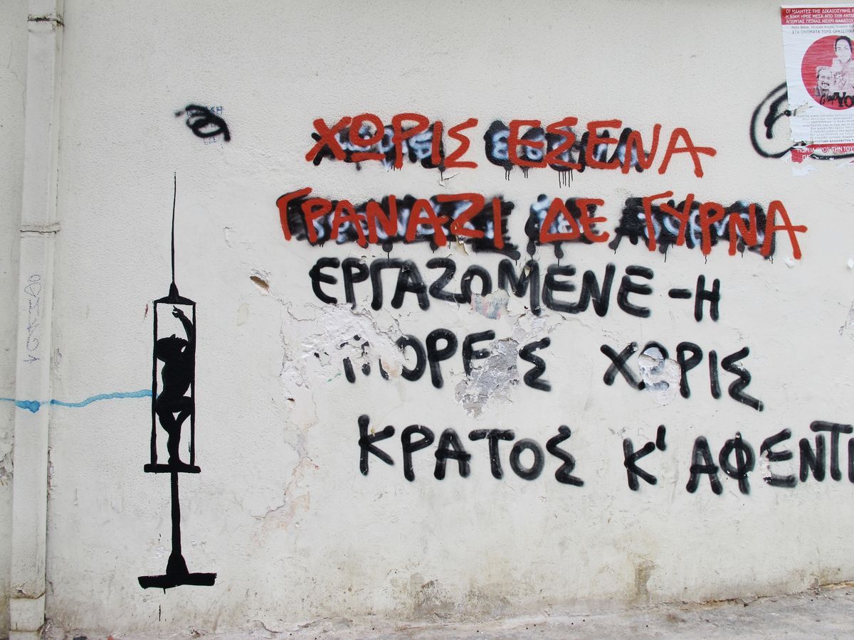 Black silhouette of a large hypodermic syringe standing upright, with a small human figure trapped inside its barrel gripping the plunger, painted beside red and black Greek graffiti text on a weathered wall.