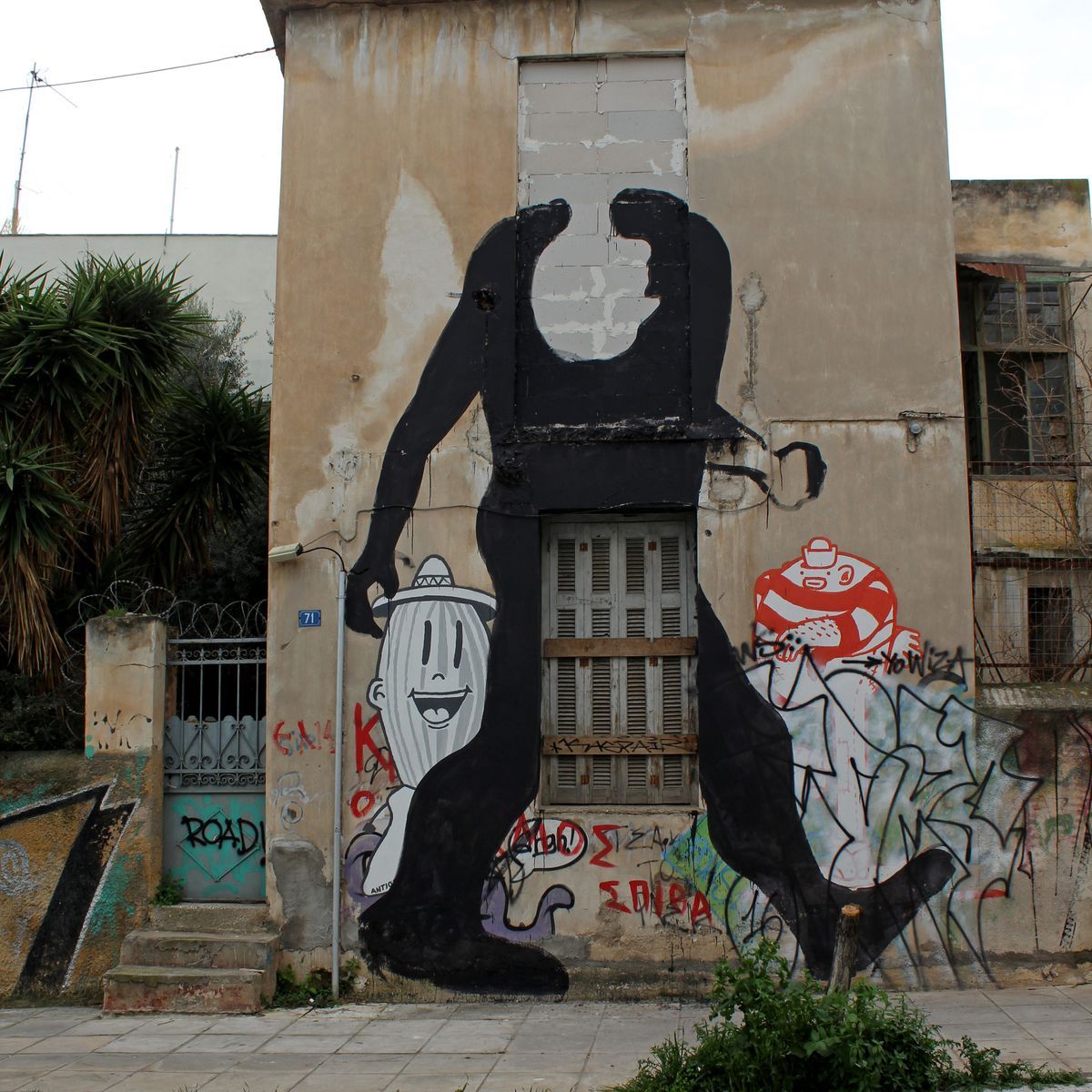 Large black silhouette of a headless, hunched humanoid figure striding across a building facade, painted beside a cartoon character wearing a hat and a red robot-like figure, over a shuttered window.