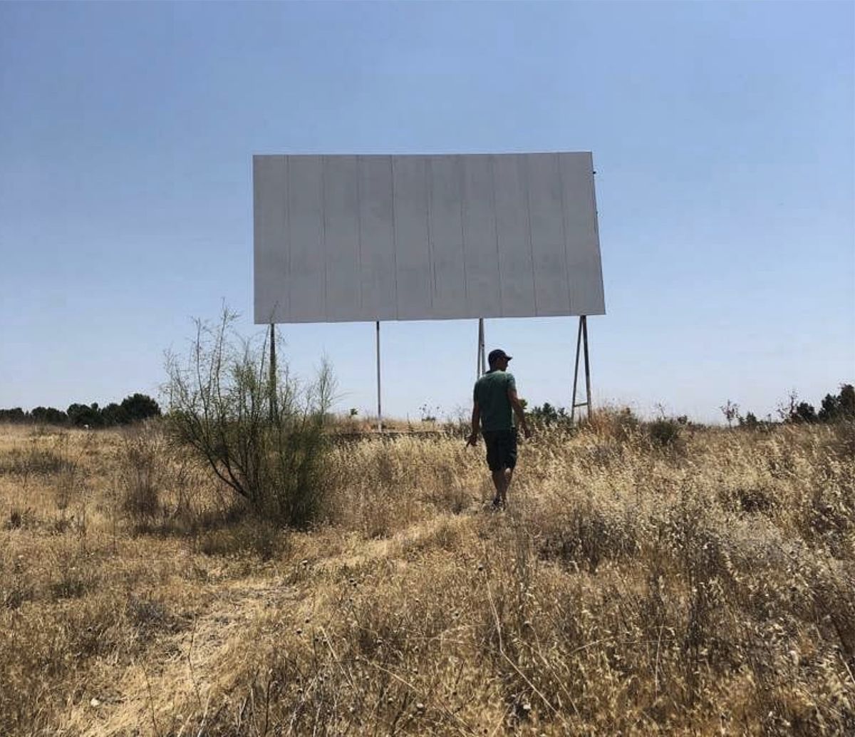 Blank white billboard mounted on metal poles in a dry grassy field, with a man walking toward it, seen from behind.