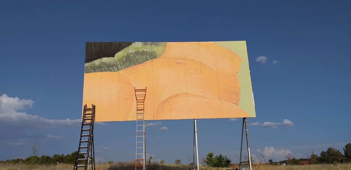 The same billboard partially painted with the first section of a mural showing a dark tree silhouette and a pale orange expanse, two ladders leaning against it, set in a dry field under a blue sky.