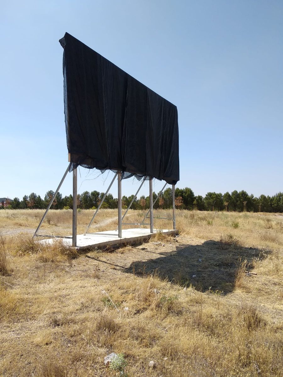 The billboard entirely covered by a black mesh fabric draped over its full width, hiding the painted homage to Courbet's L'Origine du monde, standing on its metal supports in a dry grass field with trees in the background.