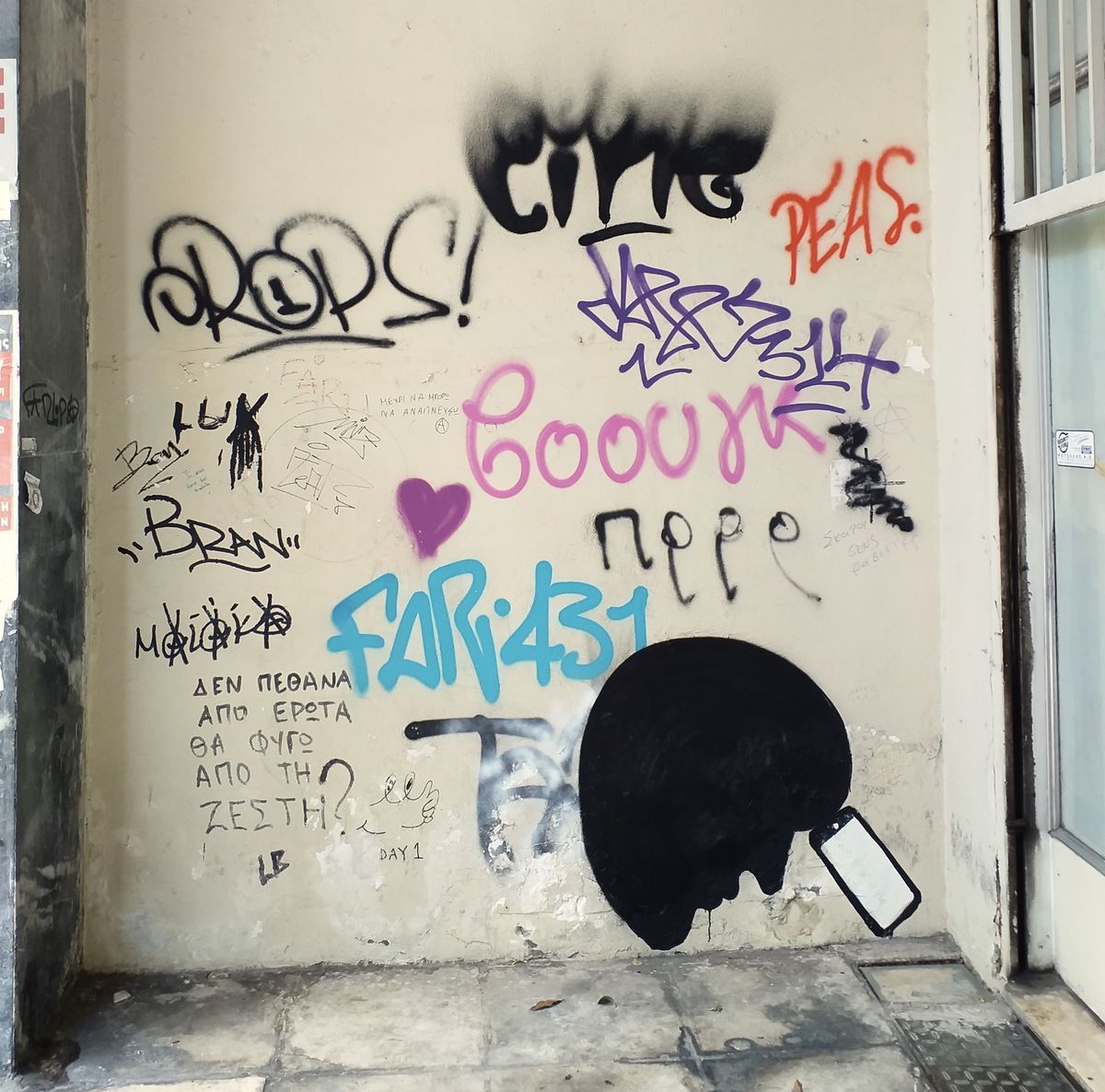 Black silhouette of a round head holding a smartphone up to its face, painted on a plastered wall covered in colorful spray-painted graffiti tags and handwritten notes.