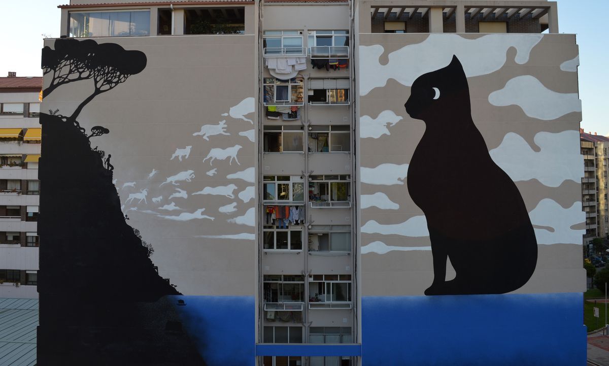 Wide view of two building facades: the left painted with a black silhouette cliff, tree, and shepherd watching a flock of white cloud-animals, the right showing the black cat with a crescent-moon eye against the camouflage-cloud pattern.