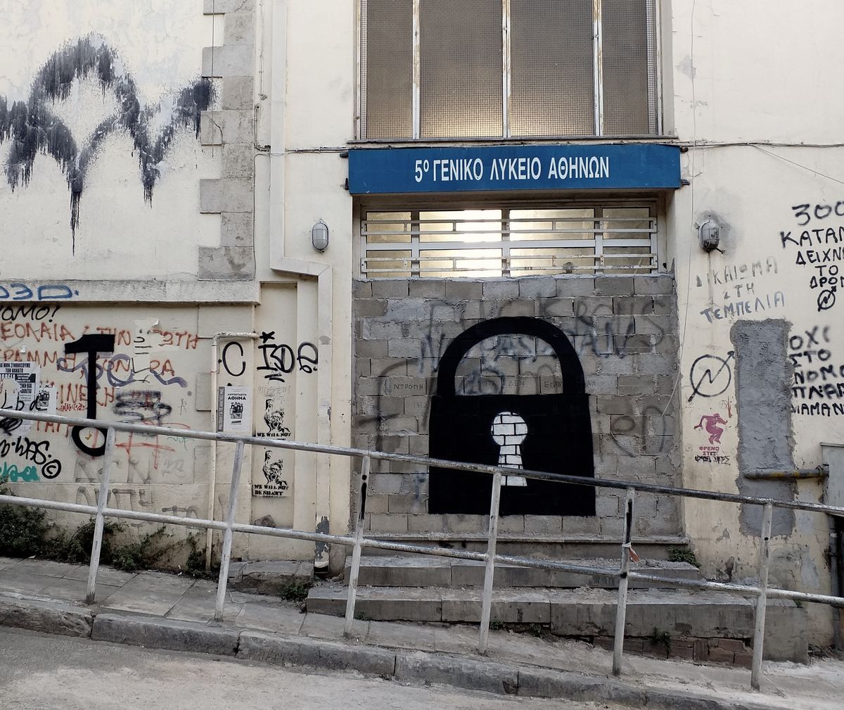 Black silhouette of a closed padlock with a white keyhole, painted over a cinder block doorway beneath a blue school name sign in Greek, flanked by metal handrails and graffiti tags.
