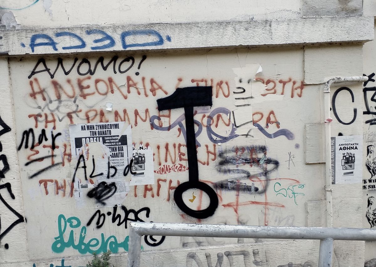 Black silhouette of an old-fashioned key painted vertically on a plastered wall covered in Greek graffiti text and layered paper flyers.