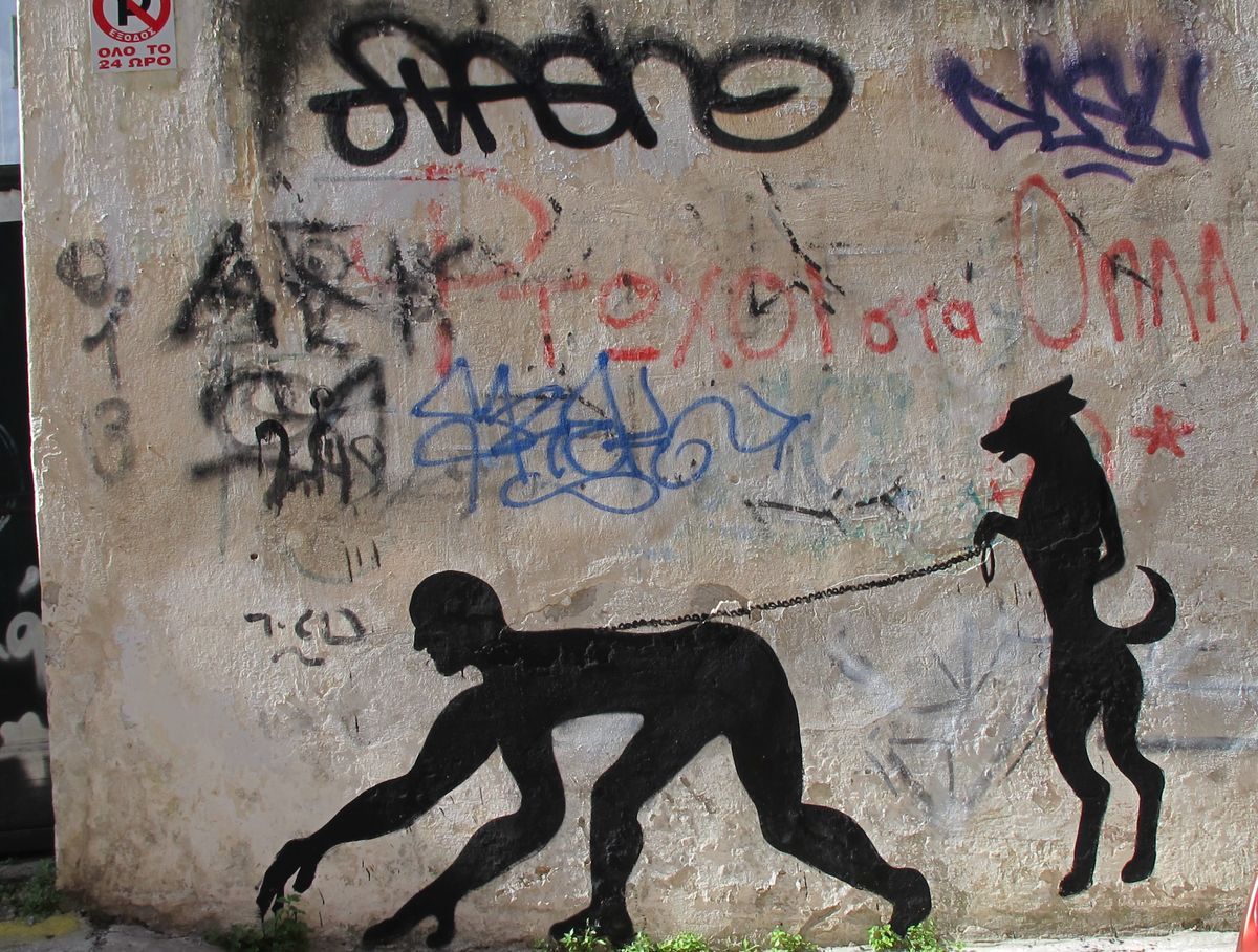 Black silhouette mural of a crouching man crawling on all fours while holding a leash attached to an upright, dog-headed humanoid figure, painted on a weathered wall covered in spray-painted graffiti.