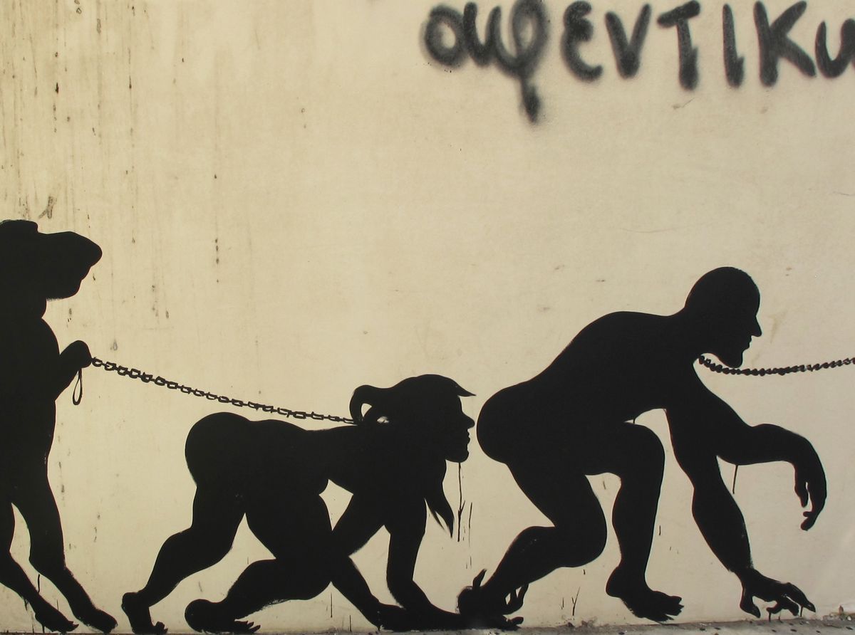 Black silhouette mural depicting an evolution-of-man sequence in which each walking figure leads the next by a chain, progressing from an animal-like form through a goat-headed figure to an upright man, painted on a plain wall beside Greek graffiti text.