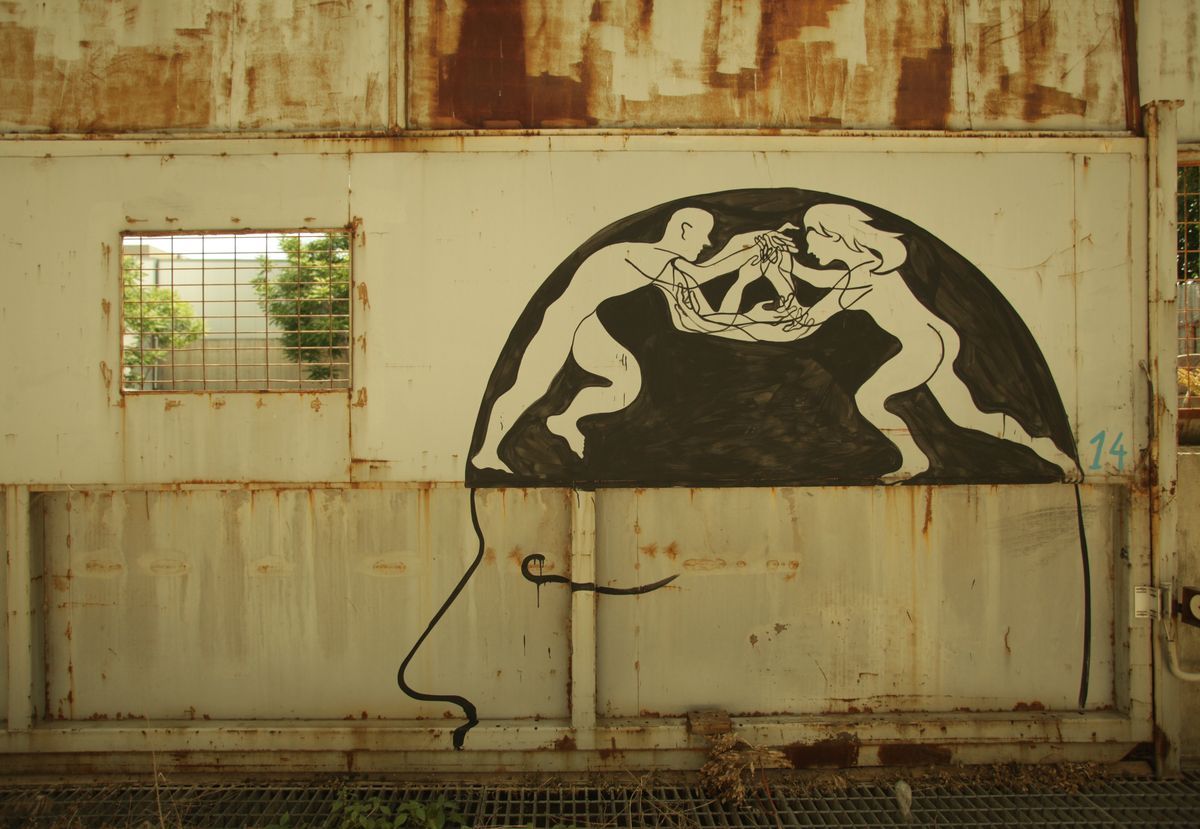 White outline mural of two nude figures pulling at a tangled mass of lines between them, painted inside a black semicircle that forms the top of a head, its profile continuing below in a thin black line down a rusted metal wall.