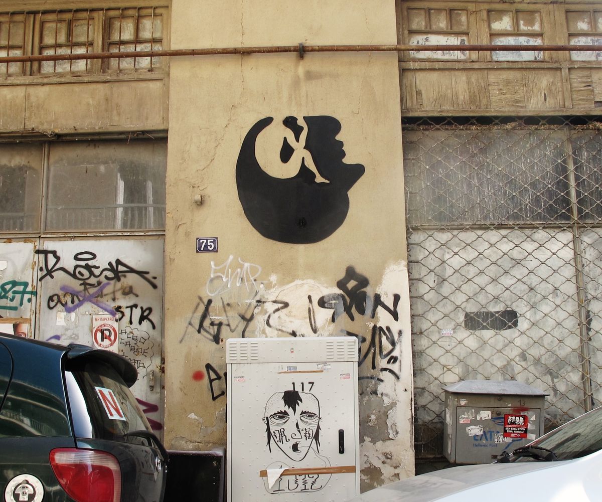 Black silhouette combining a crescent moon shape with a small curled figure inside it, merging into the profile of a human head, painted on a plastered wall beside a numbered address plaque.