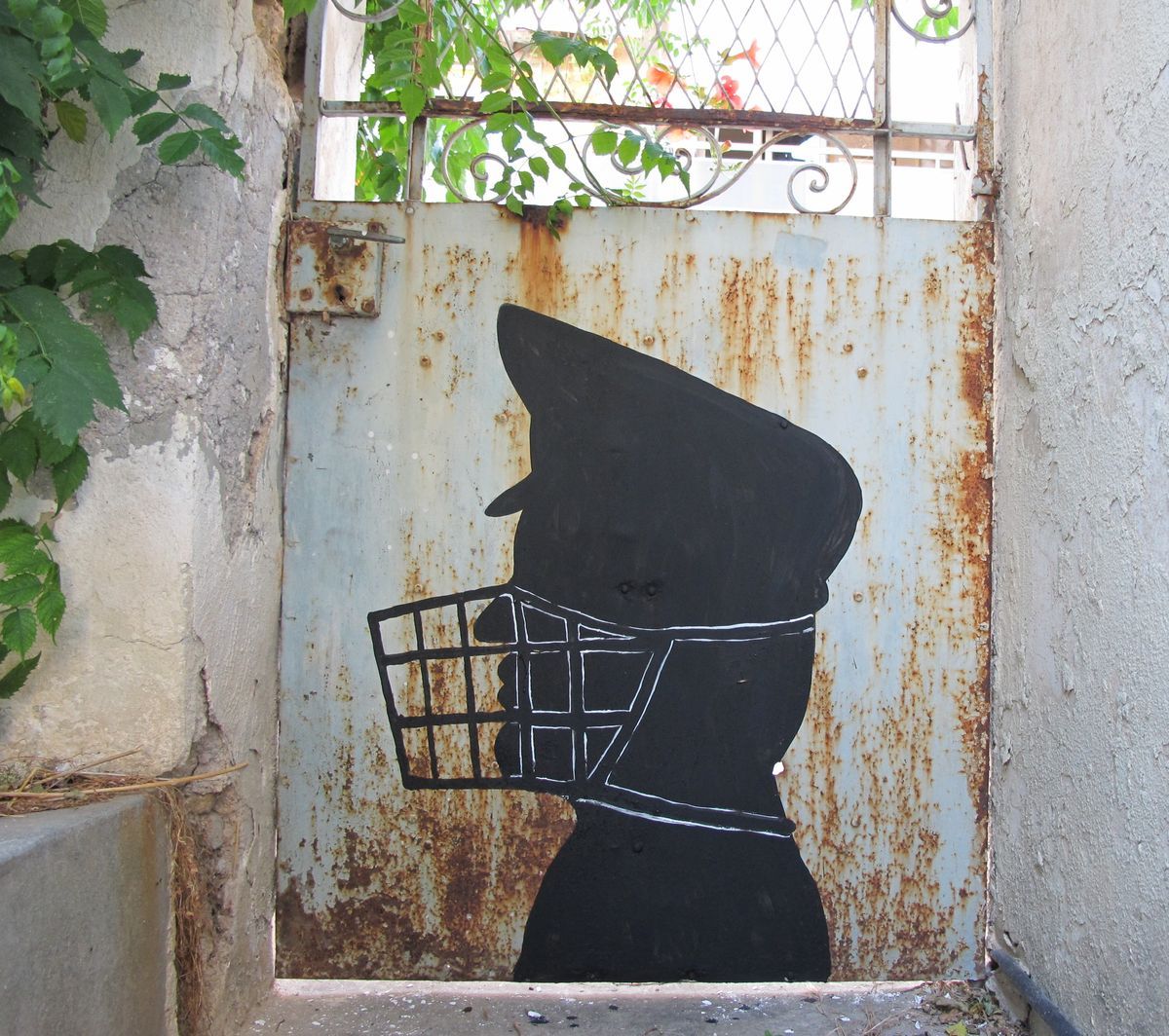 Black silhouette of a head wearing a peaked cap, its mouth covered by a white-outlined wire muzzle, painted on a rusted metal gate.