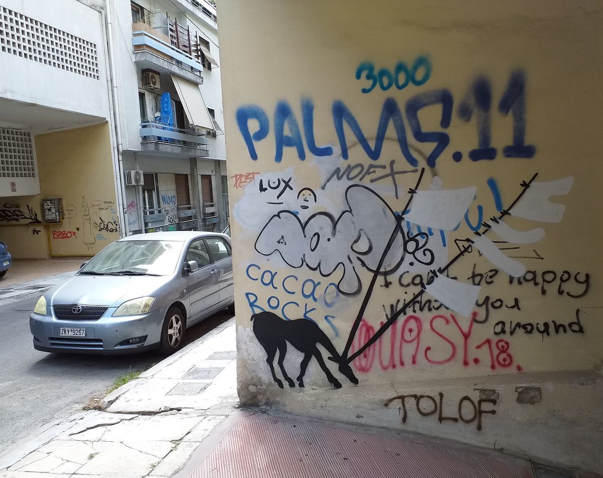 Black silhouette of a grazing goat with a diagonal clothesline strung with clothespins rising from its horns, painted on a yellow corner wall covered in spray-painted graffiti tags.