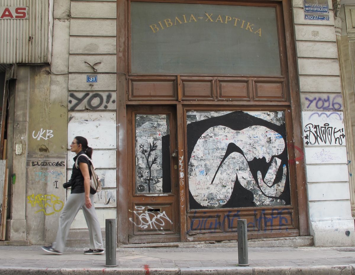 Black silhouette of a hunched, fetal-like figure formed from torn paper fragments, mounted on a glass-paneled wooden shopfront door, with a woman walking past on the sidewalk.