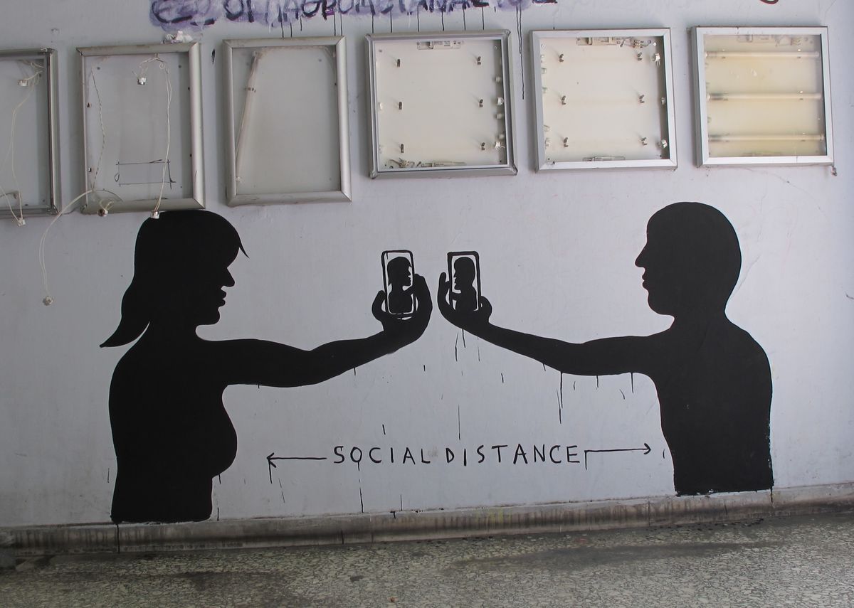 Black silhouette mural of a man and a woman standing apart, each holding up a phone showing the other's face, with the words SOCIAL DISTANCE painted between them on a white wall.