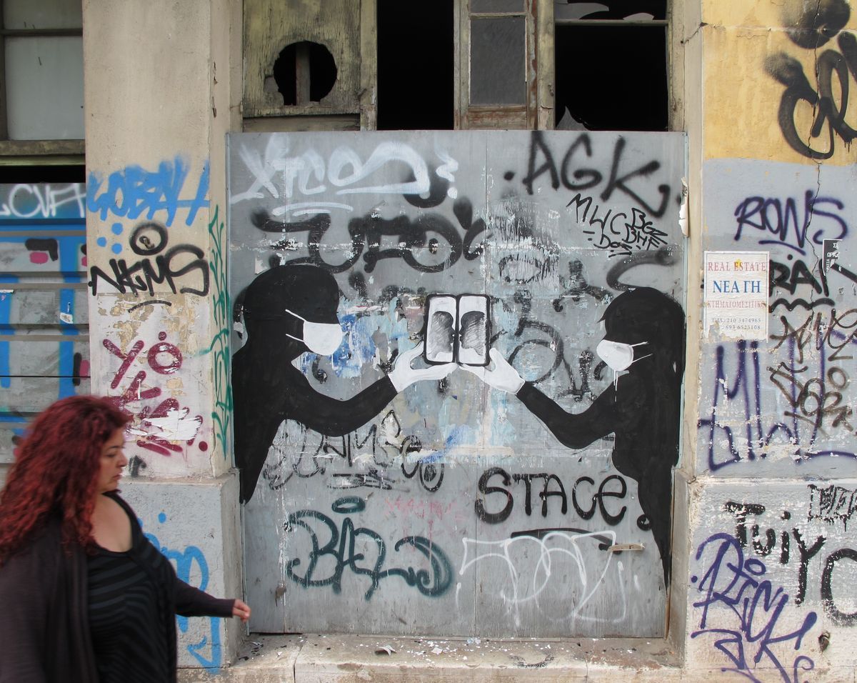 Black silhouette mural of two masked, gloved figures pressing their phones together to form a single screen, painted on a graffiti-covered wooden panel, with a passerby walking past.
