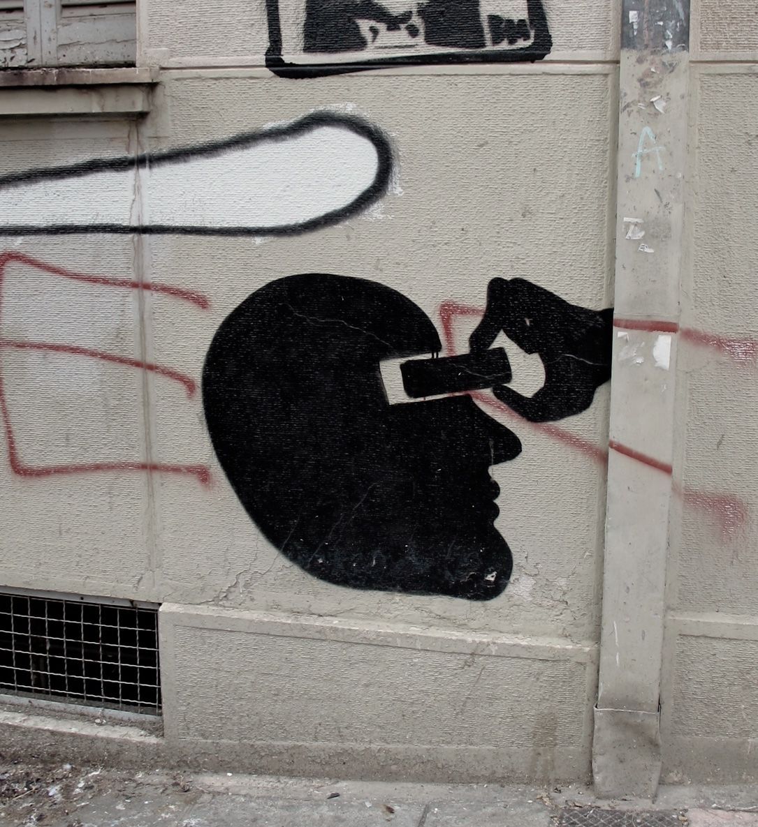 Black silhouette of a head in profile with a hand inserting a USB memory stick into a slot cut into the skull, painted on a textured wall beside red and white graffiti marks.