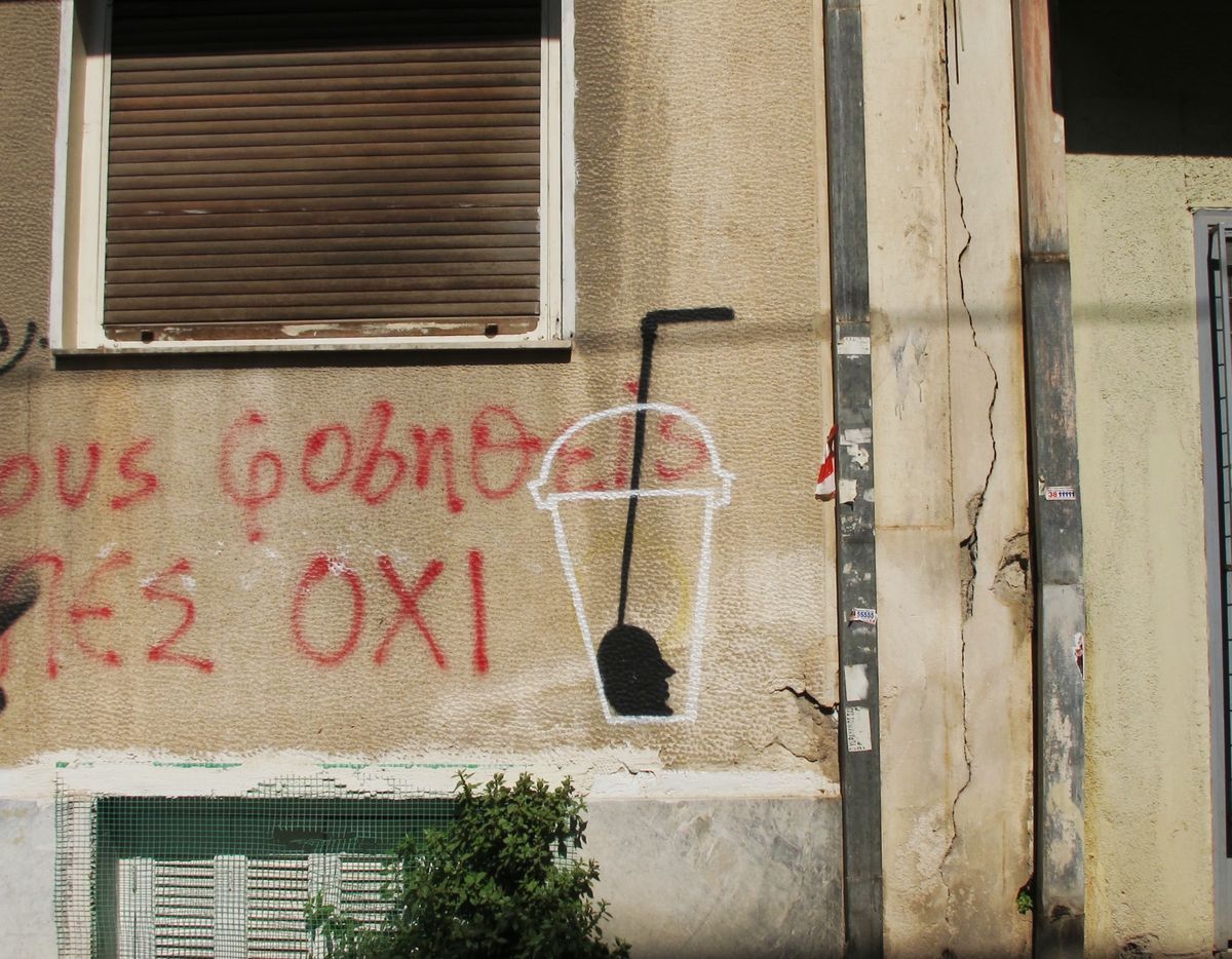 Black and white stencil of a to-go cup with a straw, its base filled in as a dark human head silhouette in profile, painted on a cracked plastered wall beside red Greek graffiti text.