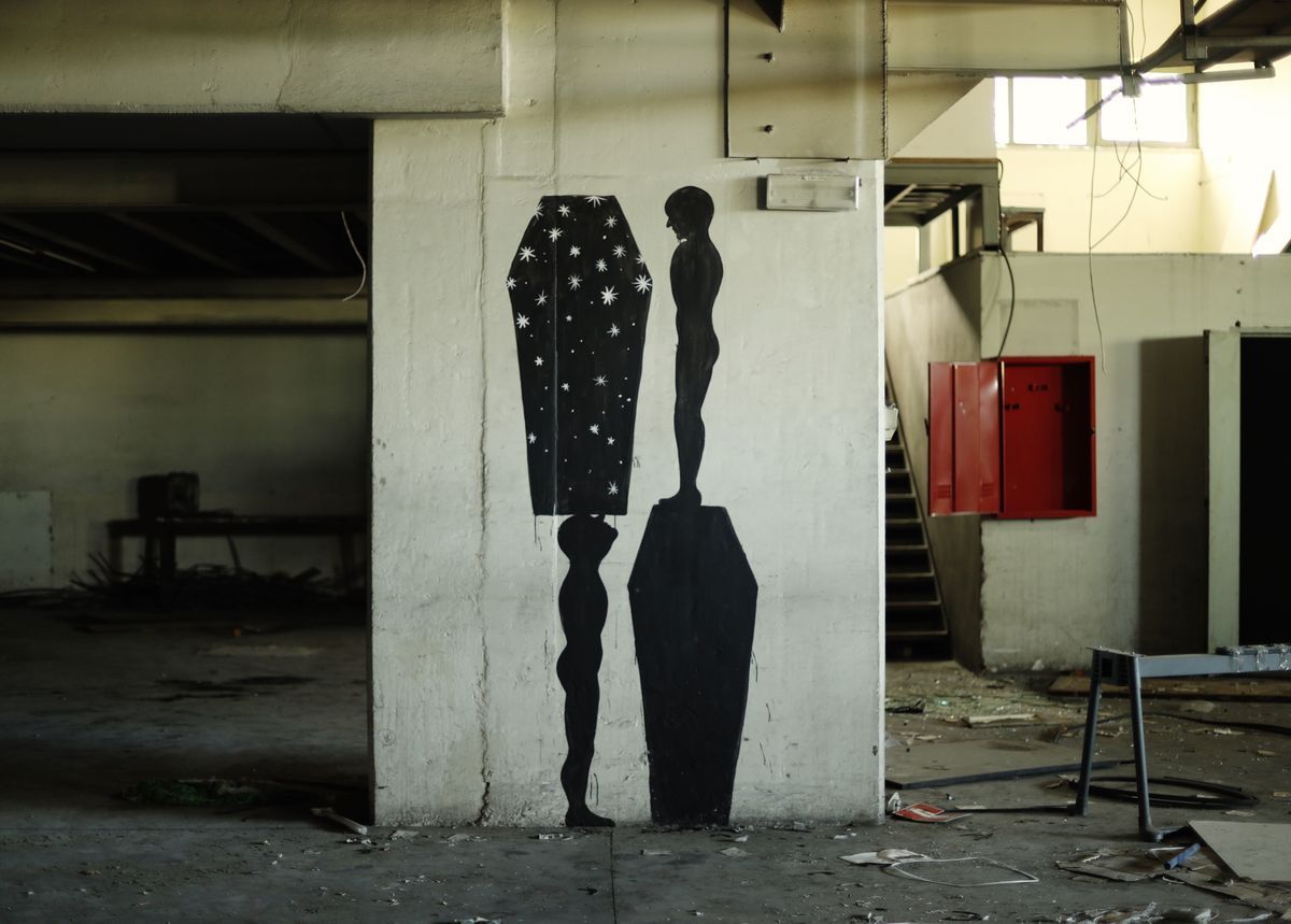 Two black silhouette figures standing on inverted podium-shaped pedestals inside an abandoned industrial building, one silhouette patterned with white stars, the other plain, surrounded by construction debris and a red electrical panel.