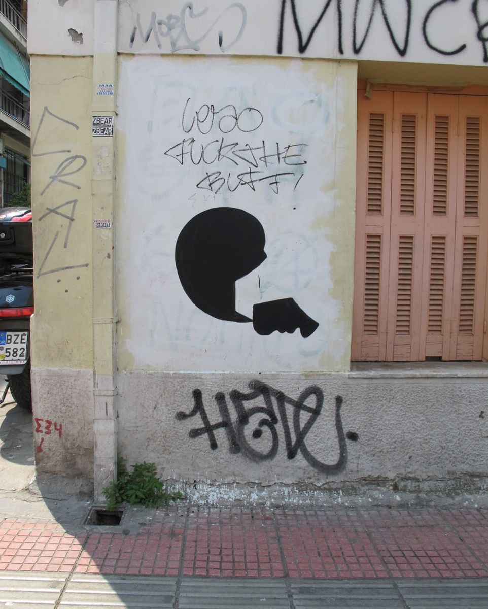 Black silhouette of a head in profile with a hand holding a face mask up to its mouth, painted on a whitewashed corner wall covered in graffiti tags, beside a pair of shuttered orange doors.