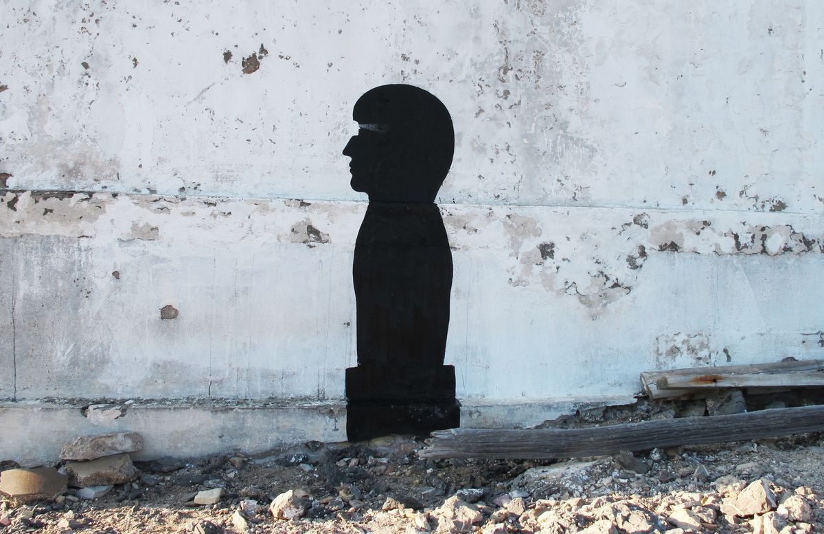 Black silhouette bust of a head and shoulders in profile, painted on a weathered white wall with peeling plaster, standing amid scattered rubble and a wooden beam on the ground.
