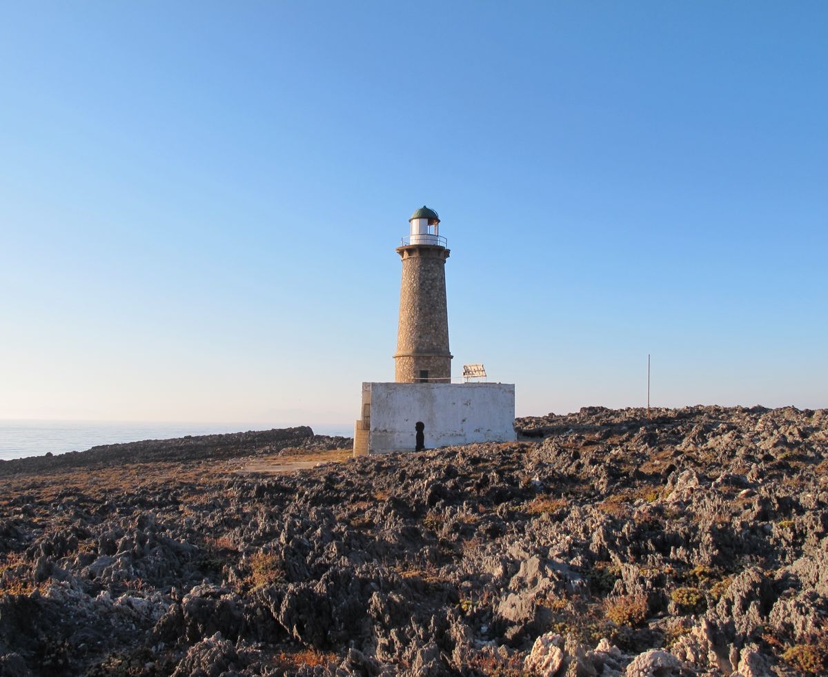 A stone lighthouse with a dark green lantern room stands beside a white outbuilding on bare rocky terrain overlooking the sea under a clear blue sky.
