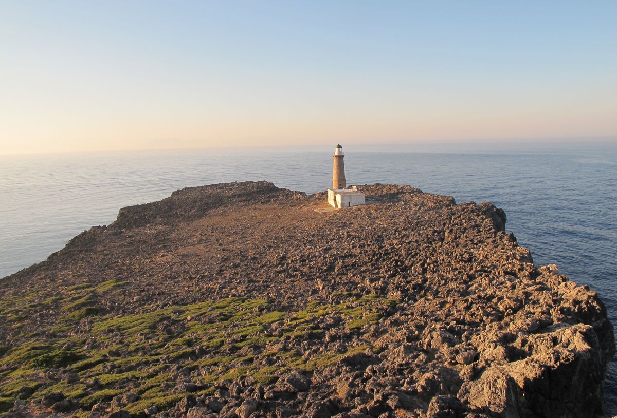 Elevated view of a rocky, sea-bound headland with a solitary lighthouse and adjoining white building at its center, lit by low golden sunlight.