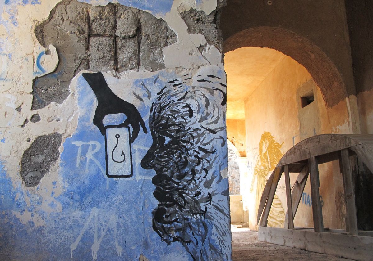 Black mural of a hand holding a fishing hook above a head rendered in loose feathered brushstrokes, painted on a peeling blue wall beside an archway leading to a golden-toned mural of a robed figure.