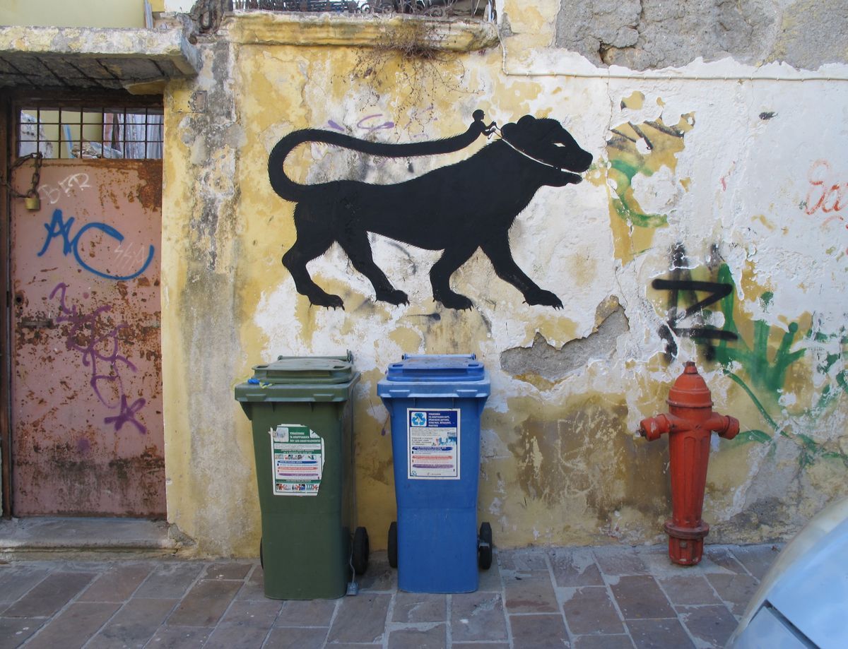 Black silhouette mural of a large dog-like animal on a leash held by a tiny figure on its back, painted above two wheelie bins and a red fire hydrant on a weathered yellow wall.
