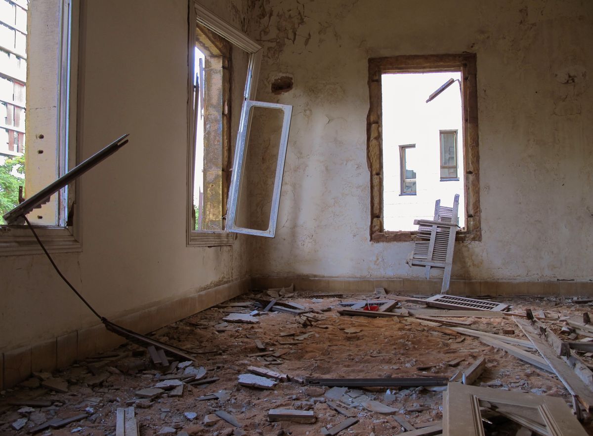 Interior of a bombed-out room with peeling walls, a mirror hanging loose from a shattered window frame, and floor debris including broken tiles and wood splinters scattered across the ground.