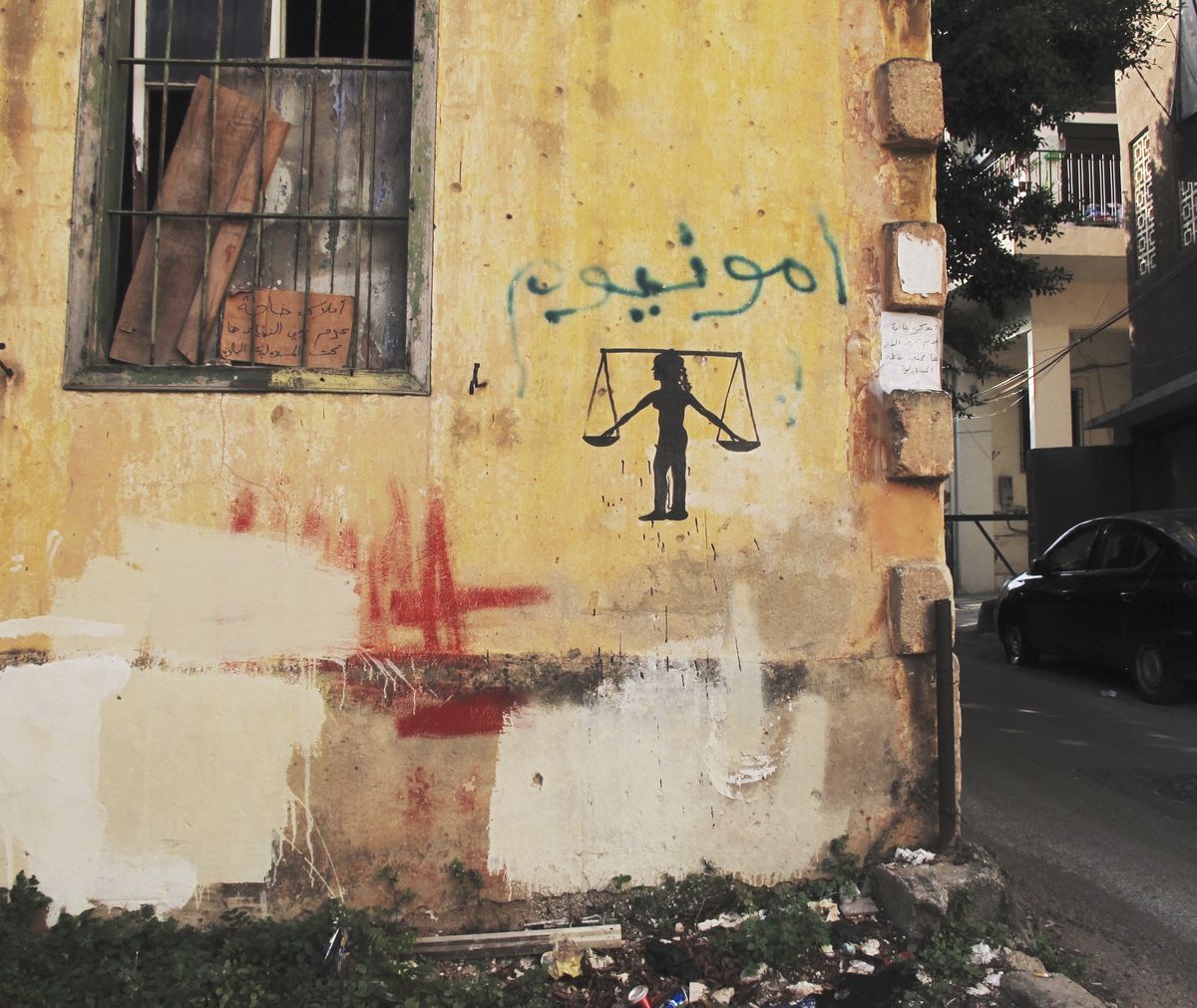 Black silhouette mural of a young figure holding a large balance scale across its shoulders, painted on a yellow wall beside Arabic graffiti text and streaks of red and white paint.