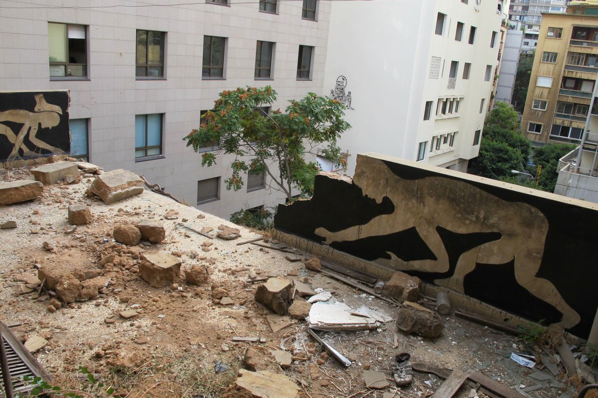 Aerial view from a rubble-covered rooftop showing two black silhouette murals of running or leaping figures painted on adjacent building walls, with a distant third silhouette visible on another building in the background.