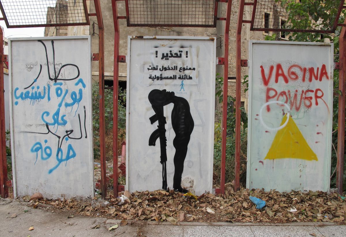 Black silhouette mural of a soldier bending down to kiss the barrel of a rifle held against the ground, painted on a metal panel between two others covered in blue and red spray-painted graffiti text.