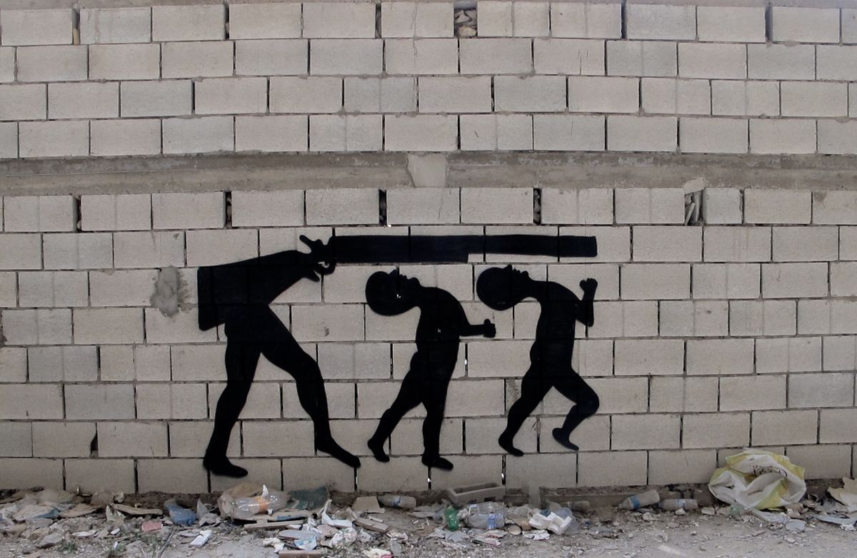 Black silhouette mural of three bent-over figures marching in a row, the front one carrying a long rifle across its shoulders, painted on a cinder block wall above scattered debris.