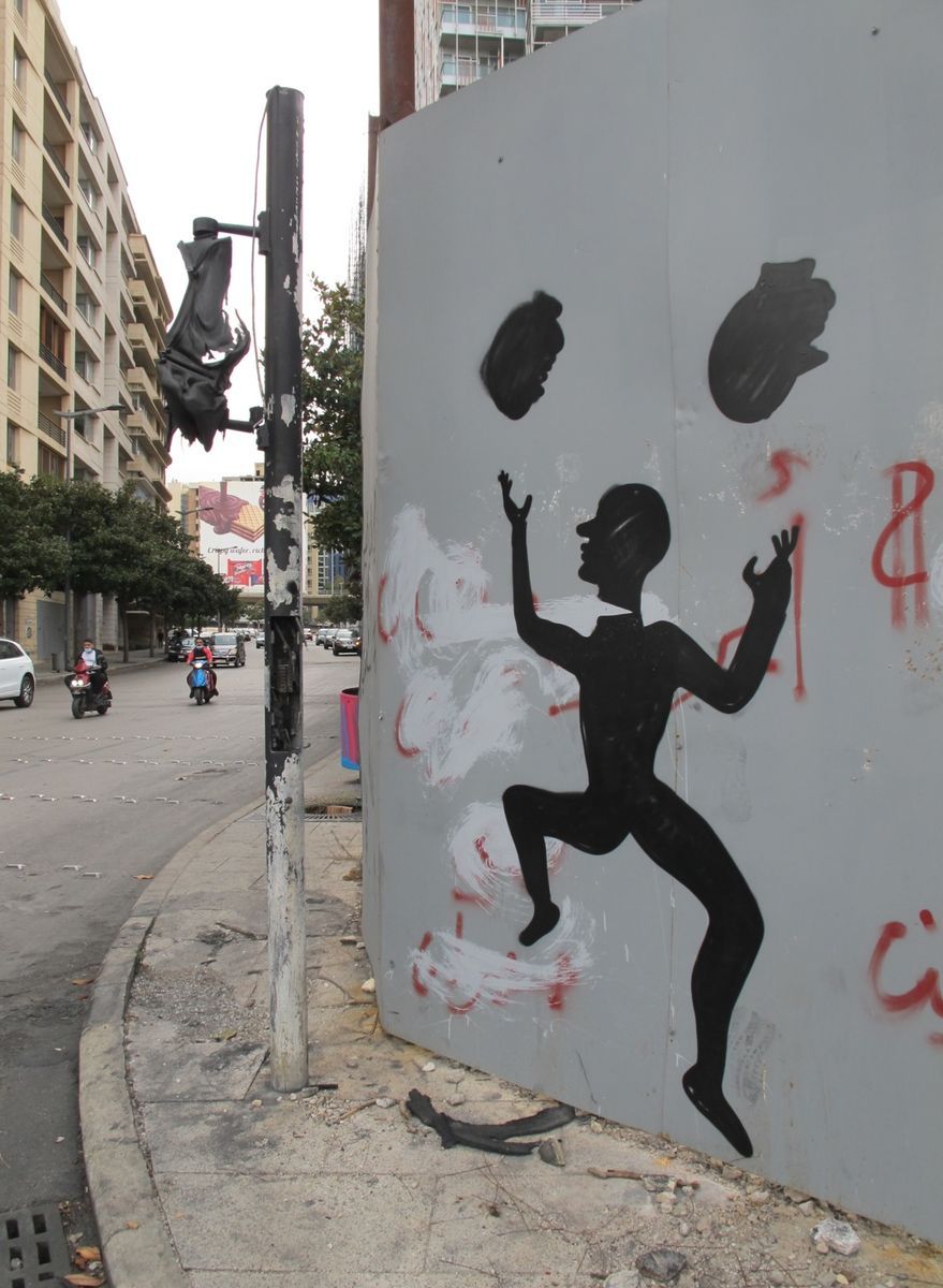 Black silhouette mural of a running figure juggling two dark round objects overhead, painted on a grey metal wall panel splashed with white and red paint, next to a city street with passing scooters.