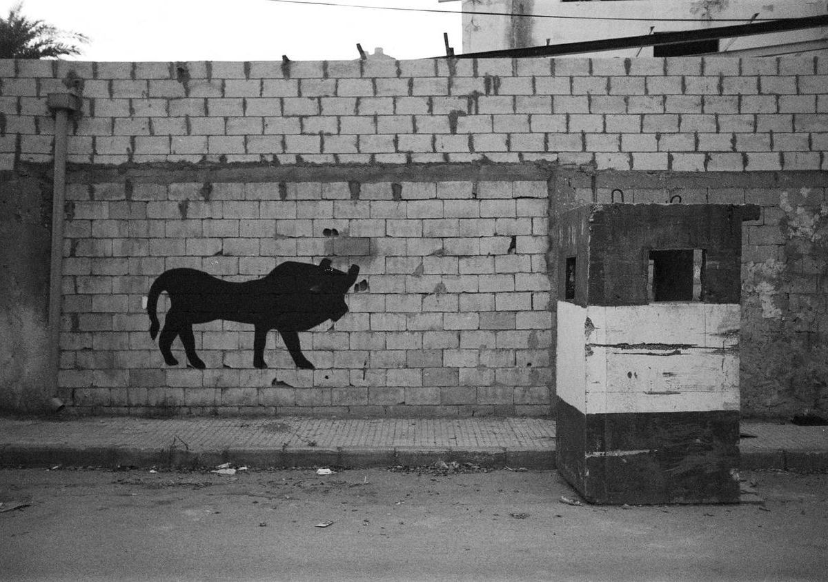 Black silhouette mural of a horned animal, resembling a bull or boar, painted on a brick wall beside a black-and-white striped guard booth.