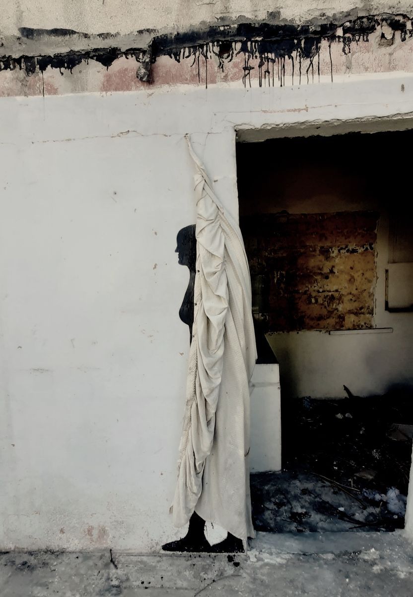 Black silhouette mural of a robed figure partially obscured by a large piece of draped cream fabric hanging beside a doorway, set against a fire-damaged wall with scorch marks and soot streaks.