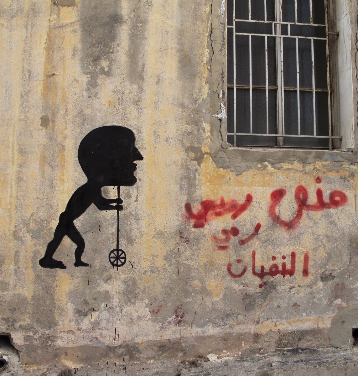 Black silhouette mural of a child figure with a large human head, bent forward while pushing a small wheeled toy on a stick, painted beside red Arabic-script graffiti on a weathered wall.