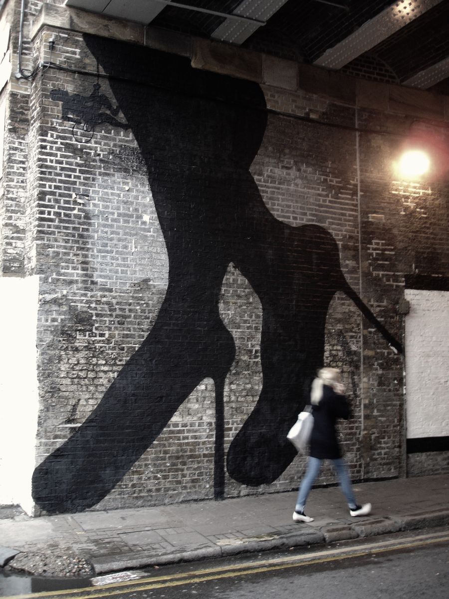 Tall black silhouette of a high-heeled leg and foot painted across a brick wall under a railway bridge, a tiny climbing figure with a rope near the top, a pedestrian walking past below.