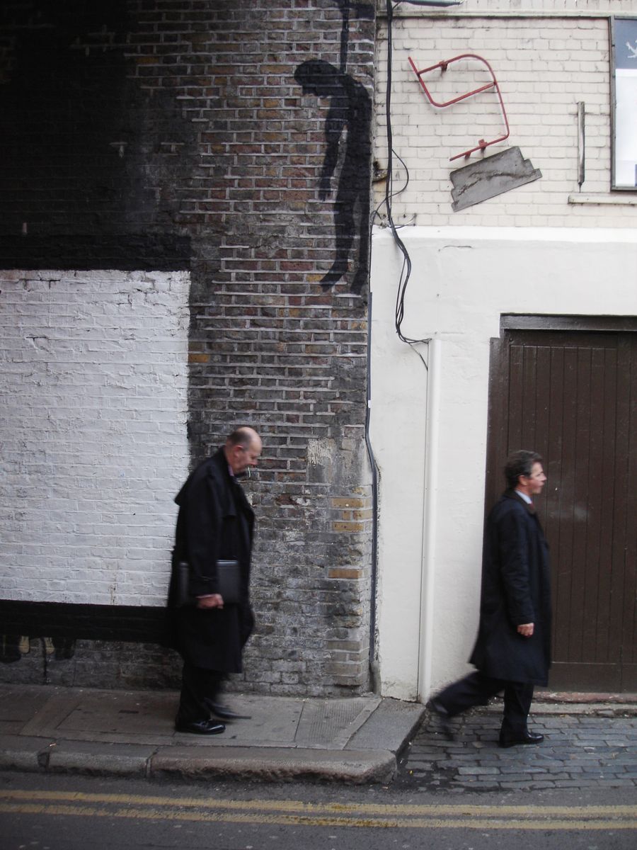 Black silhouette of a bent, downcast hanging figure painted high on a brick wall corner, two men in dark coats walking past below, red metal fixtures mounted on the wall.